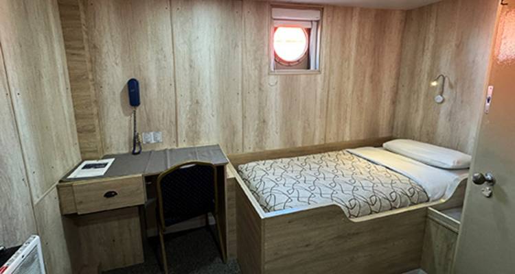 Single-berth wooden cabin with small desk and porthole providing soft daylight.