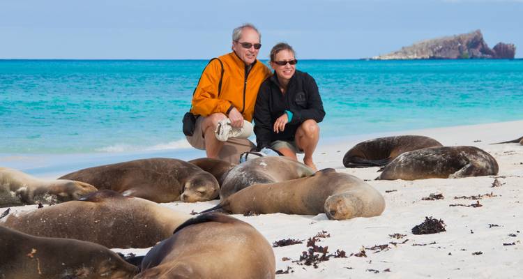 Smiling couple posing on a white beach beside sleeping sea lions and turquoise sea.