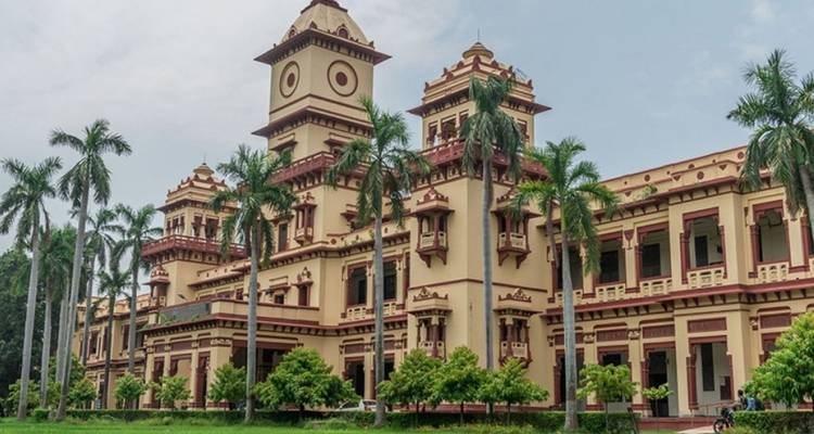Edificio institucional histórico amarillo-rojo con césped bordeado de palmeras bajo un cielo brumoso.
