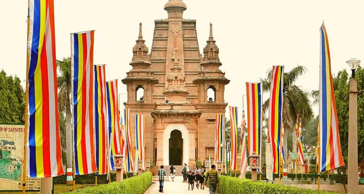 Entrada a un templo budista bordeada con banderas verticales multicolores y visitantes caminando hacia la puerta.