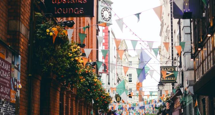 Colorful bunting and hanging flowers decorate a narrow brick street bustling with pedestrians and small shops.