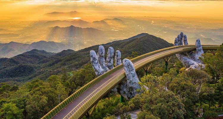 Goldene Brücke in Da Nang, gehalten von riesigen Steinhandflächen unter strahlendem Sonnenaufgangslicht über Bergketten.