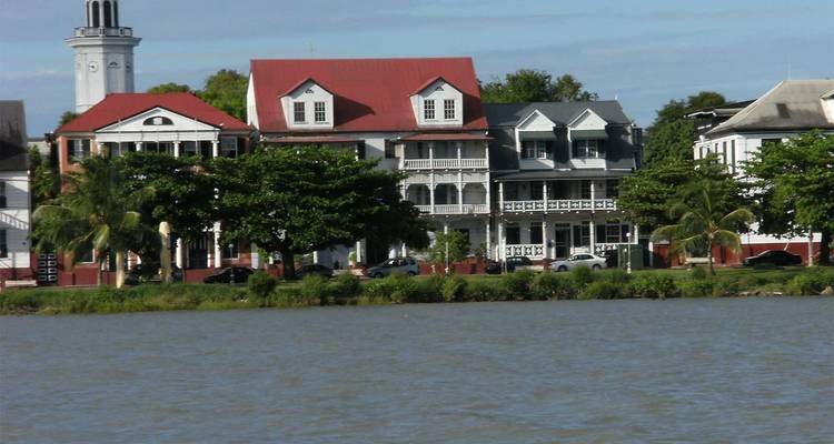 Edificios coloniales frente al mar enmarcados por árboles vistos al otro lado de un río ancho y marrón.