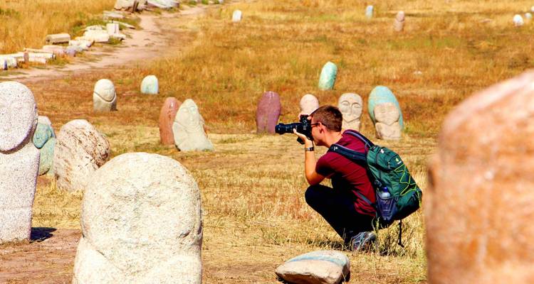 Un jeune voyageur s'accroupit avec un appareil photo pour photographier des anciennes statues de pierre balbal dispersées dans un champ herbeux aride.
