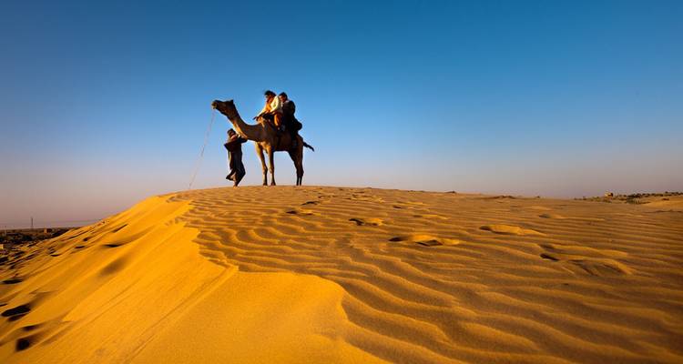Silhouettierte Kamele und Reiter überqueren eine goldene Sanddüne unter einem tiefblauen Himmel in der Wüste.