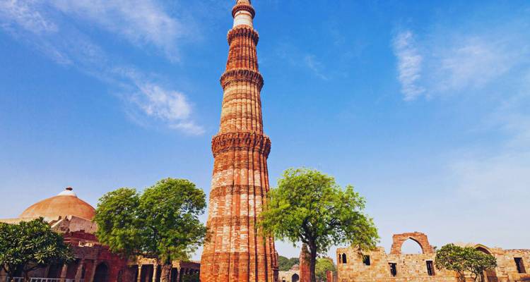 Hoher roter Sandstein-Qutub-Minar-Minarett umrahmt von strahlend blauem Himmel und belaubten Bäumen.