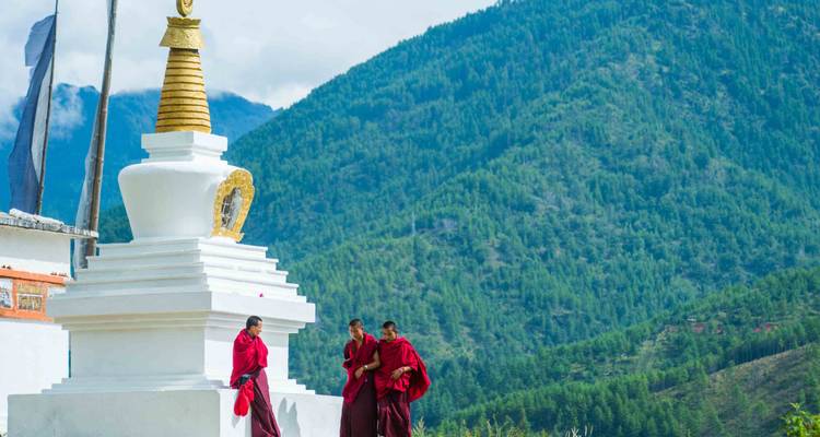 Monjes con túnicas rojas caminando junto a una estupa blanca contra las montañas boscosas del Himalaya