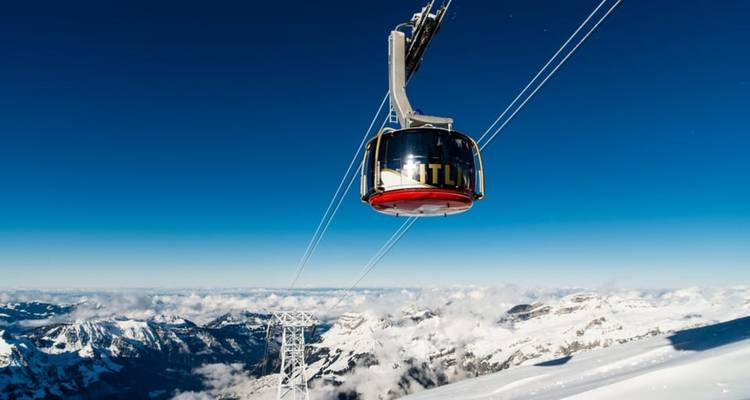 Rotating Titlis cable car glides above a sea of snowy alpine peaks and clouds under deep-blue sky.