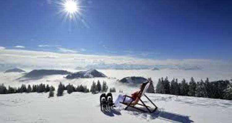 Sunlit deck chair faces a cloud-filled valley from a pristine snowfield high in the Alps.