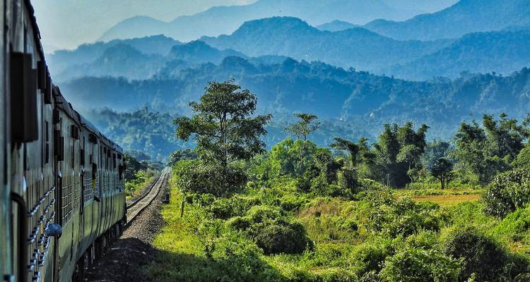 Tren de pasajeros curvando a través de un campo verde exuberante con montañas azules estratificadas en la distancia.