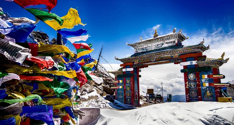 Coloridas banderas de oración ondeando junto a una puerta ornamentada de paso de montaña entre picos nevados y un cielo azul brillante.