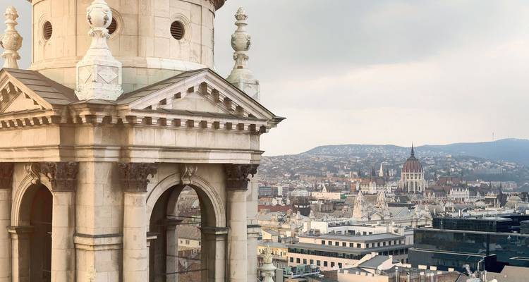 Vista cercana de la torre ornamentada de la Basílica de San Esteban con vista a los tejados de Budapest y el Parlamento en la distancia.