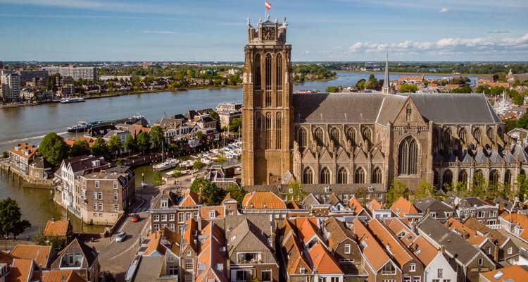 Vue aérienne de l'imposante église gothique de Dordrecht perchée au bord d'une rivière et d'un port de plaisance avec des maisons aux toits orange en contrebas.