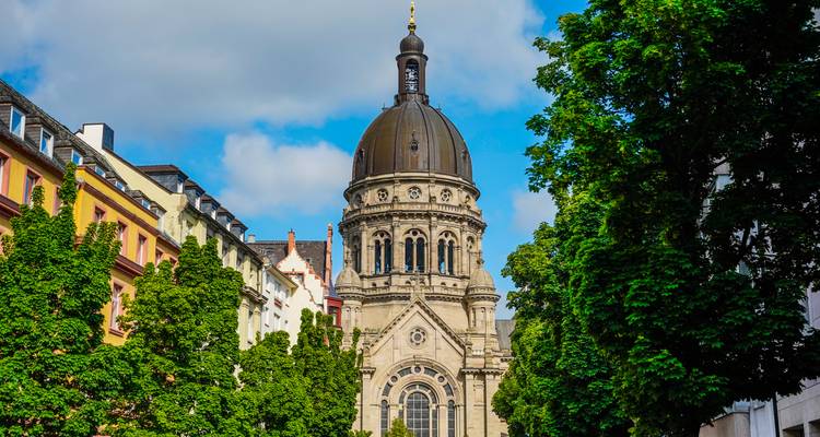 Tour d'église ornée à dôme encadrée par des arbres feuillus et des maisons de ville colorées sous un ciel partiellement nuageux