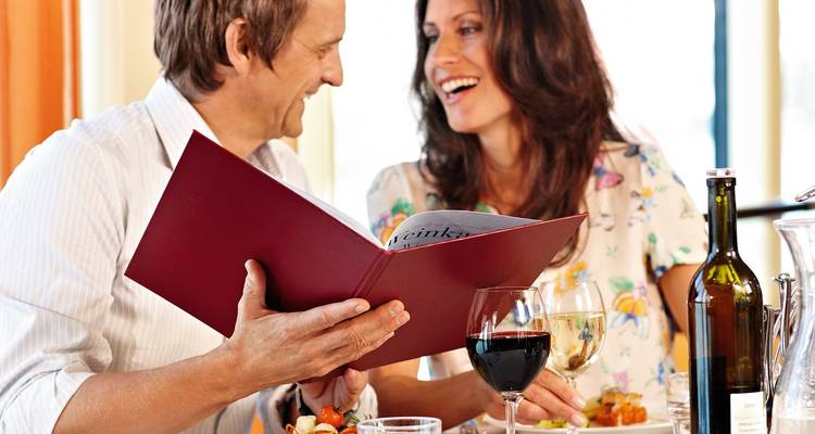 Ein lächelndes Paar genießt eine Mahlzeit in einem hellen Restaurant, der Mann hält eine geöffnete rote Speisekarte, während Weingläser und Teller mit Essen auf dem Tisch stehen.