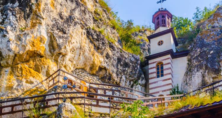 Capilla de piedra rayada del Monasterio Rupestre de Basarbovo enclavada en un acantilado escarpado con una escalera de madera que conduce hacia arriba.