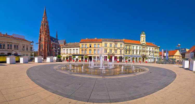 Plaza panorámica de la ciudad en Osijek con una fuente circular, fachadas históricas coloridas y la alta aguja de ladrillo rojo de la catedral.