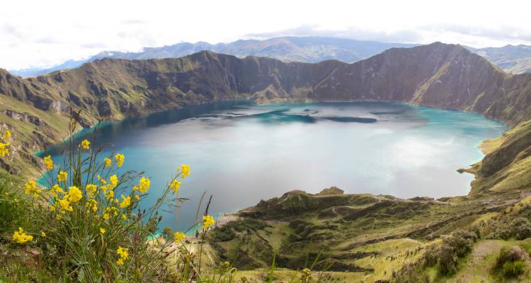 Panoramablick auf den türkisfarbenen Quilotoa-Kratersee, umgeben von schroffen Andengipfeln und Wildblumen.