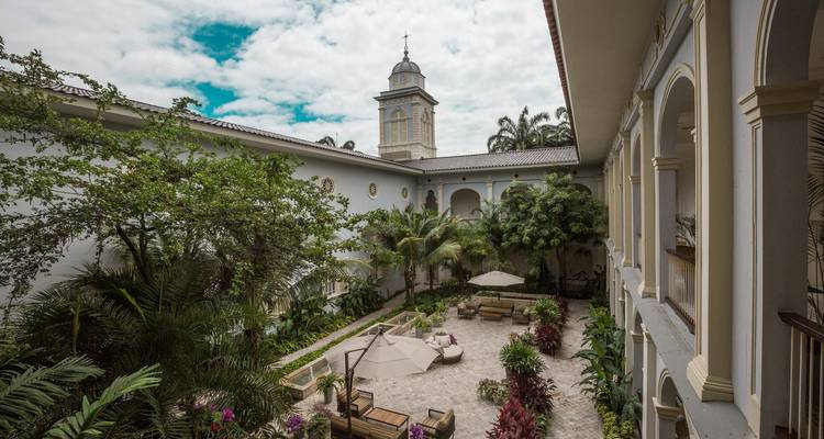 Ruhiger kolonialer Innenhofgarten mit tropischen Pflanzen und weißen Bögen unter einem teilweise bewölkten Himmel.