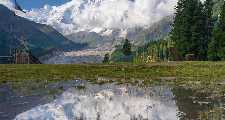 Sommet himalayen enneigé parfaitement reflété dans une flaque calme sur une plaine herbeuse encadrée de grands conifères.