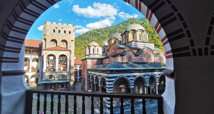 Vista vibrante de la iglesia rayada y la torre del Monasterio de Rila enmarcadas por un arco con fondo de montañas verdes.