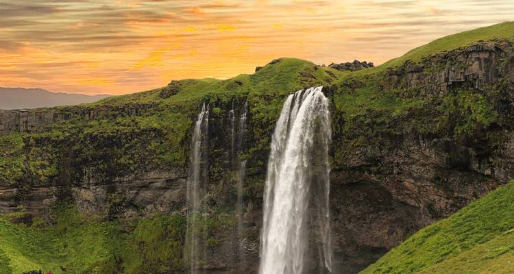 Seljalandsfoss stürzt über grüne Klippen unter einem warmen pastellfarbenen Himmel hinab.