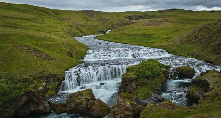 Ein sanfter stufenförmiger Wasserfall fließt durch ein üppig grünes isländisches Tal.