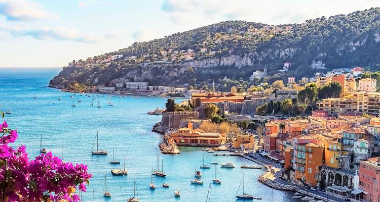 Lebendige Aussicht über die azurblaue Bucht und die farbenfrohen Küstenhäuser der Französischen Riviera vor bewaldeten Hügeln mit verstreuten Villen.
