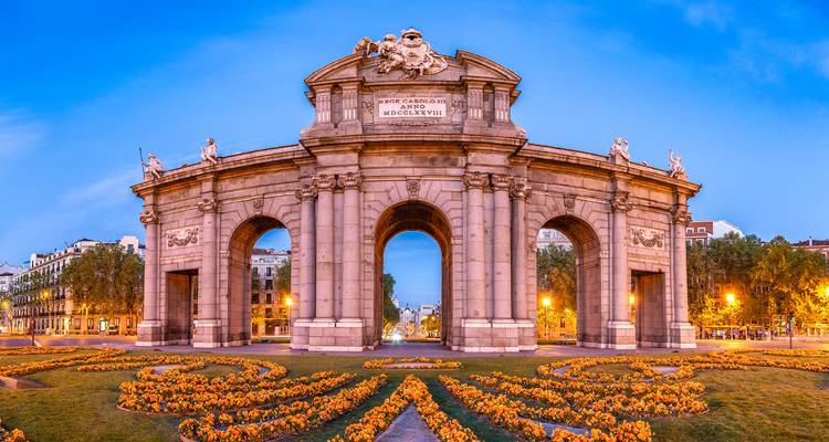 Monumento de la Puerta de Alcalá en Madrid iluminado al atardecer con vibrantes parterres de flores en primer plano.