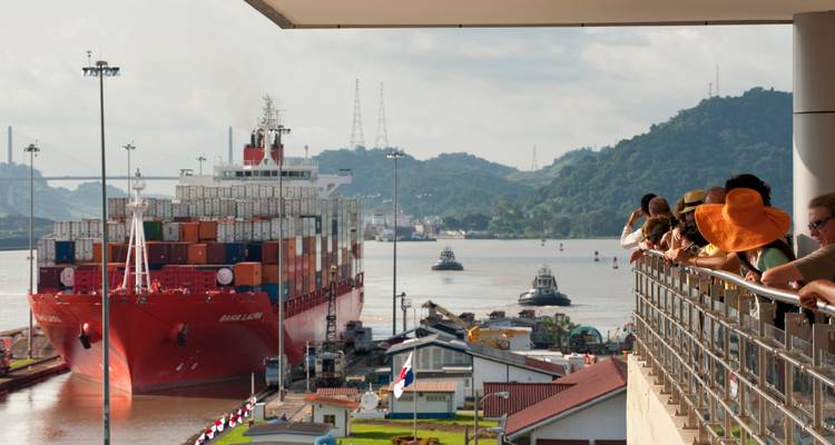 Spectators watch a large container ship transit the Panama Canal locks with lush hills in the background.