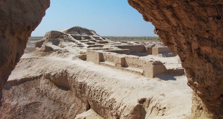 Durch eine felsige Öffnung erstrecken sich sonnengebackene Erdwände und Fundamente einer alten Zitadelle in die flache Wüste.