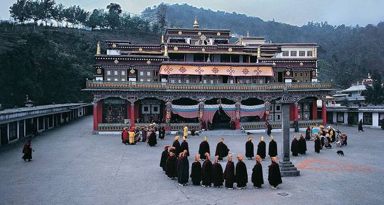 Monks in maroon robes assemble in the large courtyard of an ornate hilltop monastery complex.