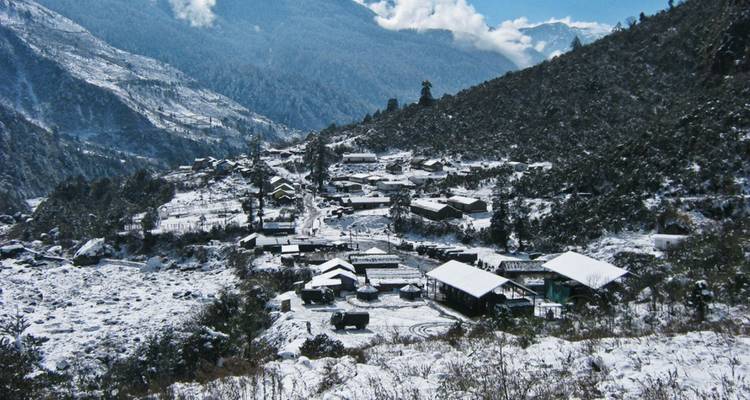Snow-dusted mountain village viewed from above, surrounded by dark evergreen forest.