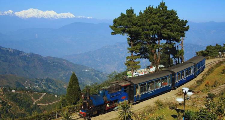 Vintage blue steam locomotive pulling carriages along a mountainside with panoramic Himalayan peaks in the distance.