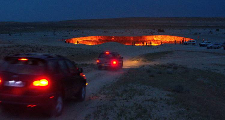 Escena nocturna de vehículos conduciendo hacia el ardiente cráter de gas de Darvaza que brilla en el desierto.