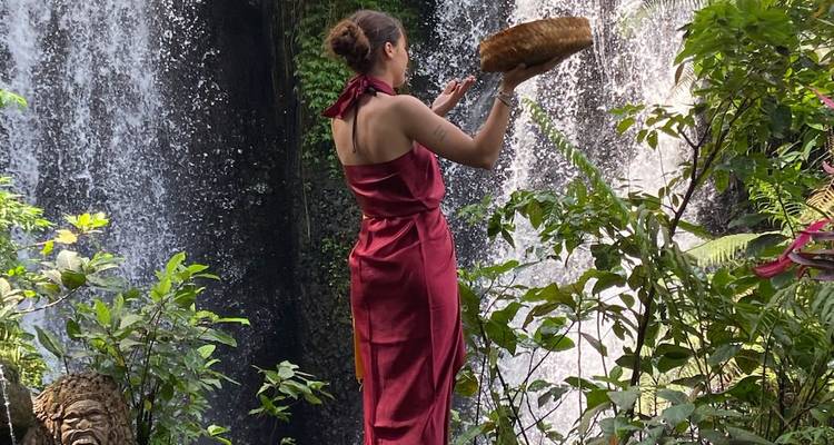 Femme en tenue de cérémonie offre un panier près d'une cascade tropicale entourée de verdure.