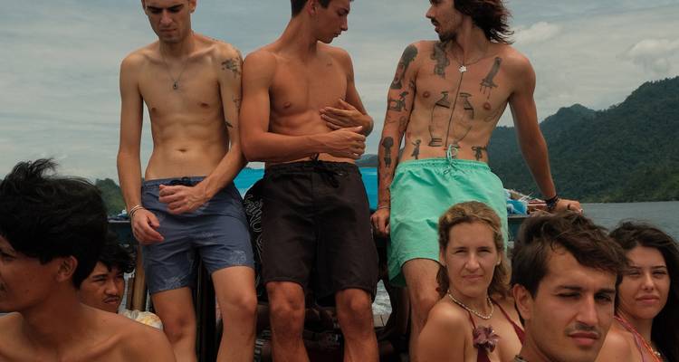 Des voyageurs se tiennent sur le pont d'un bateau en mouvement, les cheveux dans le vent avec des collines vertes derrière.