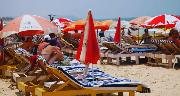Escena de playa tropical abarrotada con bañistas en tumbonas de madera bajo sombrillas coloridas cerca del mar.
