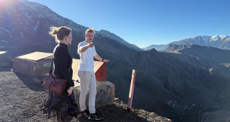 Reisende, die auf einem hohen Bergpass stehen und auf schroffe Atlasgipfel unter klarem Himmel blicken.