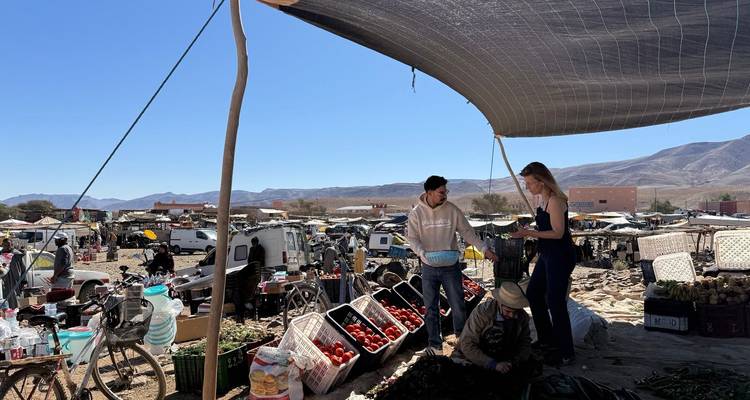 Freiluftmarkt im ländlichen Marokko mit Kisten voller Erzeugnisse unter einer Plane vor Wüstenhügeln.