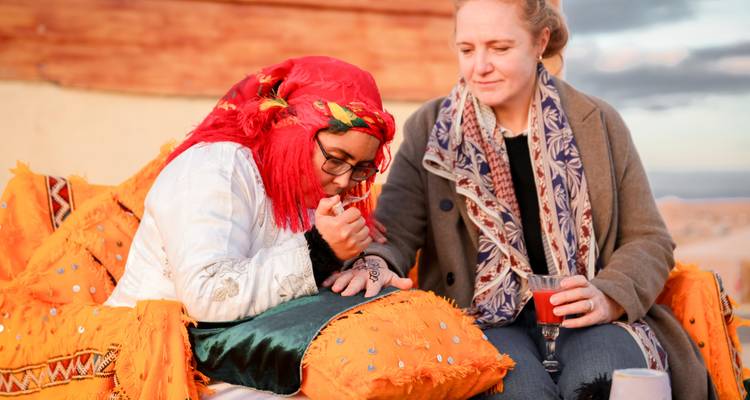 Zwei Frauen entspannen auf orangen Kissen, eine probiert Tee in einem Wüstencamp