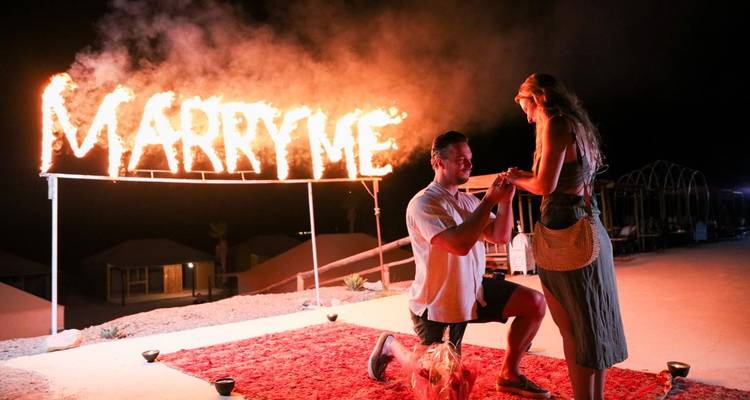 Romantischer nächtlicher Heiratsantrag mit brennendem „HEIRATE MICH"-Schild, während ein Partner kniet