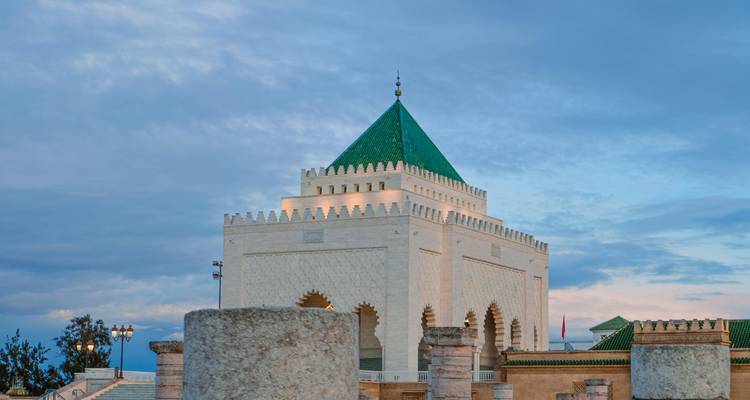 Weißes Marmormausoleum mit grünem pyramidenförmigem Dach vor sanftem Abendhimmel, eingerahmt von steinigem Vordergrund