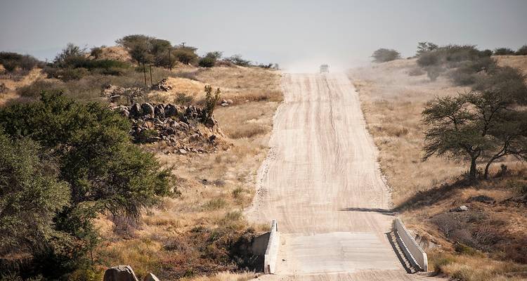 Stoffige gravelweg strekt zich uit over glooiende savanne richting veraf voertuig onder wazig Namibisch lucht.