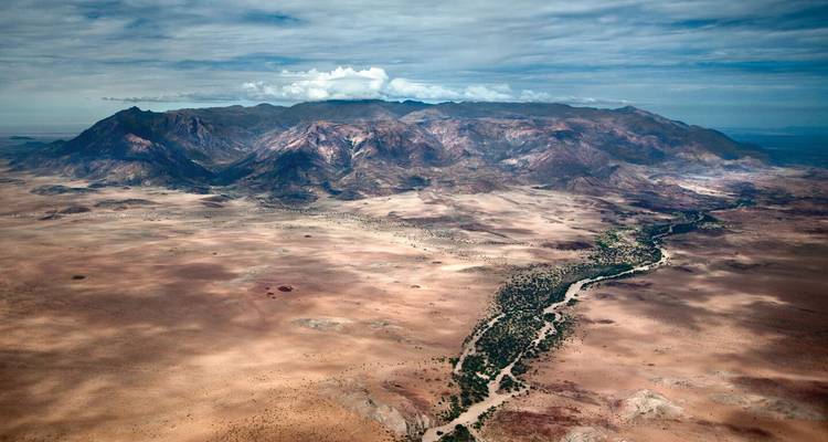 Luchtfoto van het uitgestrekte, dorre Brandberg-massief dat oprijst boven eindeloze woestijnvlaktes met een droge rivier bedding.