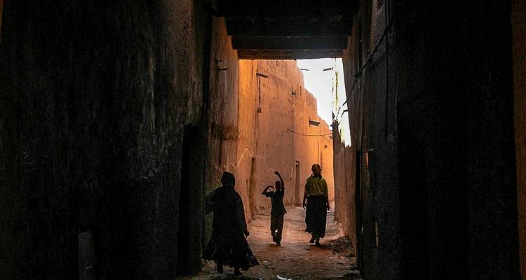 Silhouetten von Frauen und einem Kind gehen durch eine schmale, schattige Gasse, die von warmem Abendlicht in einer alten Medina beleuchtet wird.