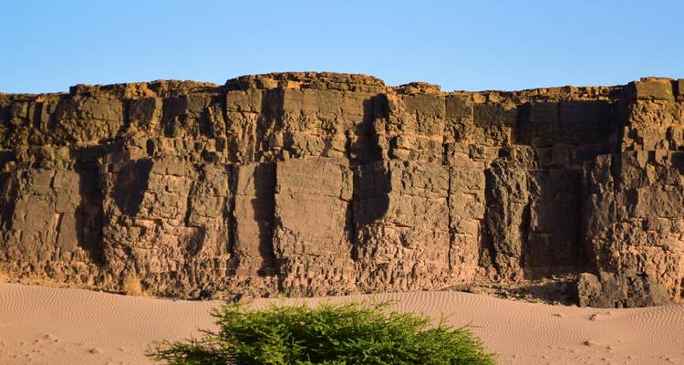 Schroffe Vulkanstein-Klippe, die sich über sandige Wüste erhebt, mit einem einzelnen grünen Strauch an ihrem Fuß und klarem blauen Himmel.