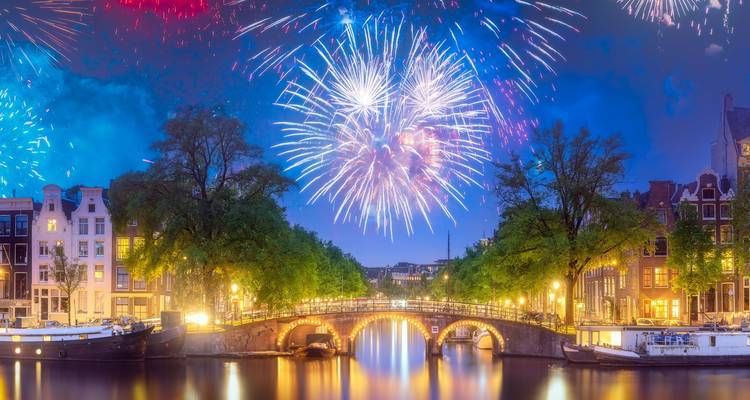 Amsterdam-Kanal bei Nacht beleuchtet mit buntem Feuerwerk, das über einer gewölbten Brücke explodiert.