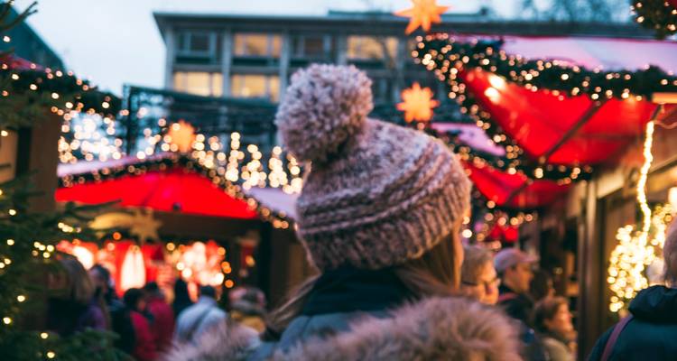 Rückansicht einer Person mit Wollmütze, die festliche Weihnachtsmarktlichter und -stände bewundert.