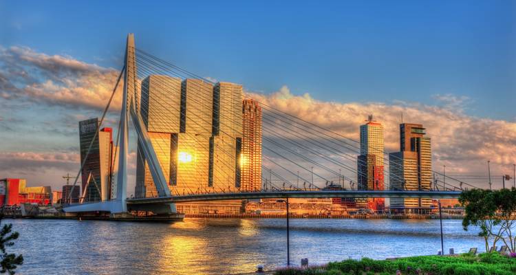Moderne Rotterdamer Skyline mit Erasmusbrücke und gläsernen Wolkenkratzern, die im warmen Licht des Sonnenuntergangs leuchten.